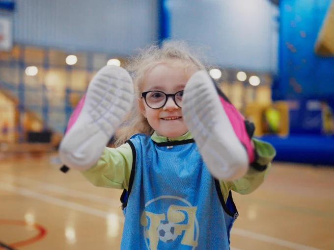 A person with glasses and long hair is smiling while holding up both feet, wearing pink-soled shoes, on an indoor sports court. 