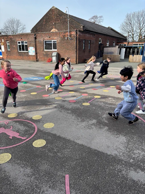 Children are joyfully playing a game on colourful markings in a schoolyard. The background features a brick building and trees under a clear sky.