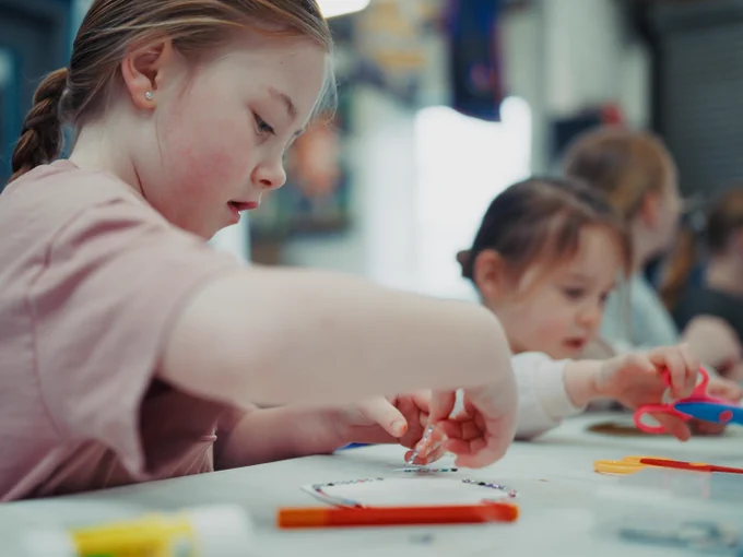 Children are sitting at a table, engaged in a craft activity using colourful materials like paper and markers. The atmosphere appears creative and focused.