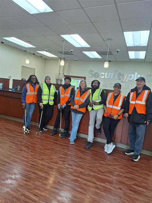 A group of seven people wearing orange safety vests pose inside a credit union. The mood is collaborative and professional.