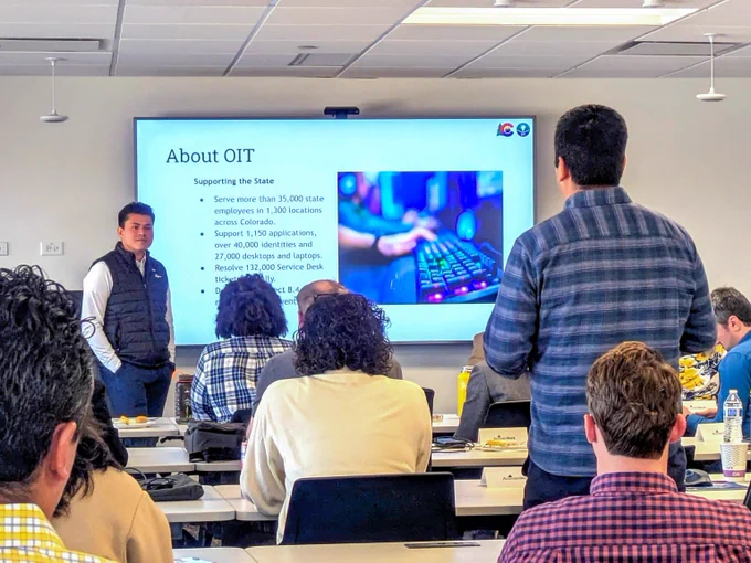 A presenter listens to a question from an audience member in a modern classroom with a large screen displaying information about OIT's services.