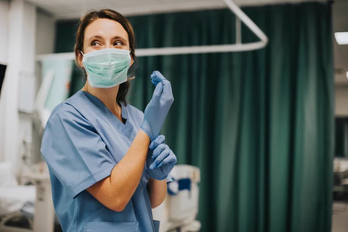 Imagen de una enfermera, que viste una mascarilla, poniéndose unos guantes de látex en un hospital (copyright: Rawpixel.com-Adobe Stock).