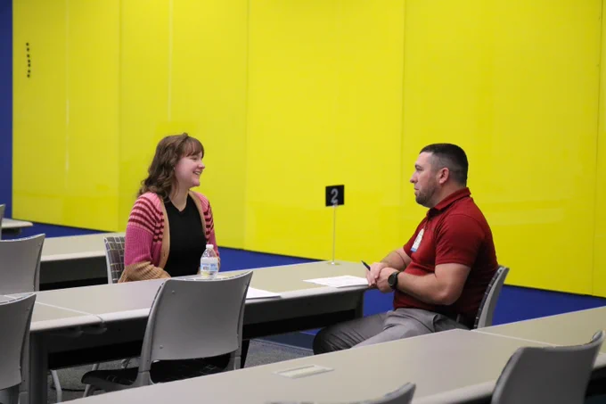 Two people seated across from each other at a table in a brightly lit room with yellow walls, engaged in conversation.