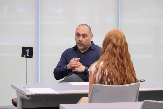 A man and a woman seated across a table in a formal interview setting with a number one sign on the table.
