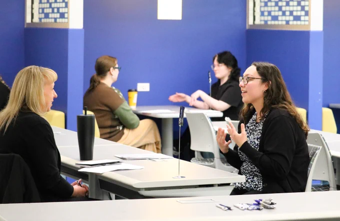 Four women engaged in a discussion while seated at tables in a room with blue walls and patterned tiles.