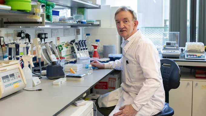 Researcher Sir Adrian Hill seated at a lab workstation surrounded by pipettes, test tubes, and electronic equipment in a bright lab.