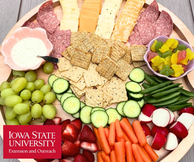 Round platter with assorted cheeses, salami, crackers, cucumber slices, green grapes, strawberries, baby carrots, radishes, green beans, and two bowls of dip and gummy candies