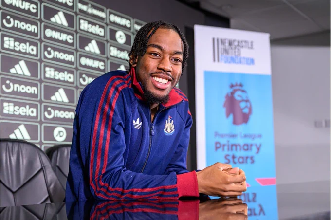 Anthony Elanga a Newcastle United jacket smiles while sitting at a press conference table. Behind them are banners displaying the Premier League Primary Stars logo and Newcastle United Foundation branding.