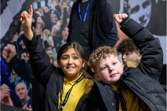 Two children wearing yellow shirts and black jackets are raising their hands in front of a mural featuring a crowd. The background shows several figures, possibly fans celebrating.