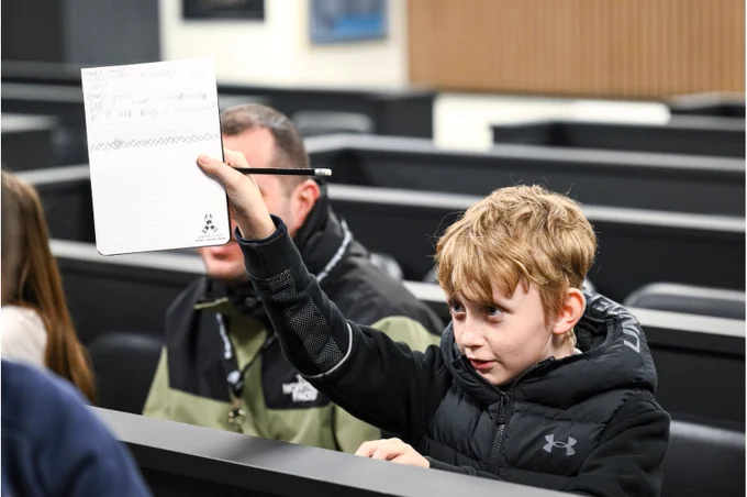 A young individual raises a notebook with writing on it, sitting in a room with several others. The person wears a dark Under Armour jacket and holds a pencil. The atmosphere appears to be in an educational or presentation setting.
