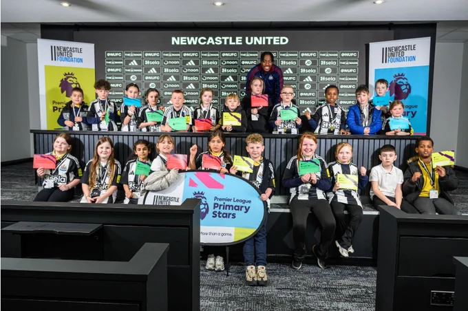 A group of children poses with certificates in a press room setting, featuring Newcastle United branding and Premier League Primary Stars logos. They are seated and standing in front of a backdrop, with some holding banners and others wearing medals. The room is branded with Premier League and Newcastle United Foundation elements.