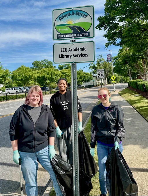 Three people are standing on a sidewalk holding trash bags and grabbers, participating in a street cleaning activity. They are positioned near a sign that reads "Adopt-A-Street, Keep Greenville Beautiful," and below it, "ECU Academic Library Services." The weather is sunny with clear skies, and trees line the street in the background.