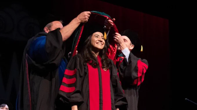 Woman smiling in academic attire being hooded by two individuals also in academic attire