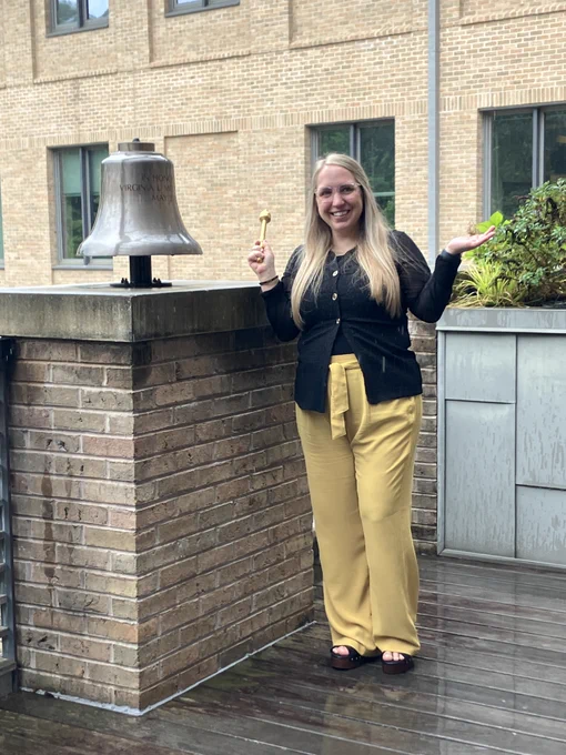 Person standing outdoors beside a mounted bell on a brick pedestal, holding a small mallet toward the bell. The setting appears to be outside a campus building with brick walls and windows, suggesting a ceremonial or milestone moment.
