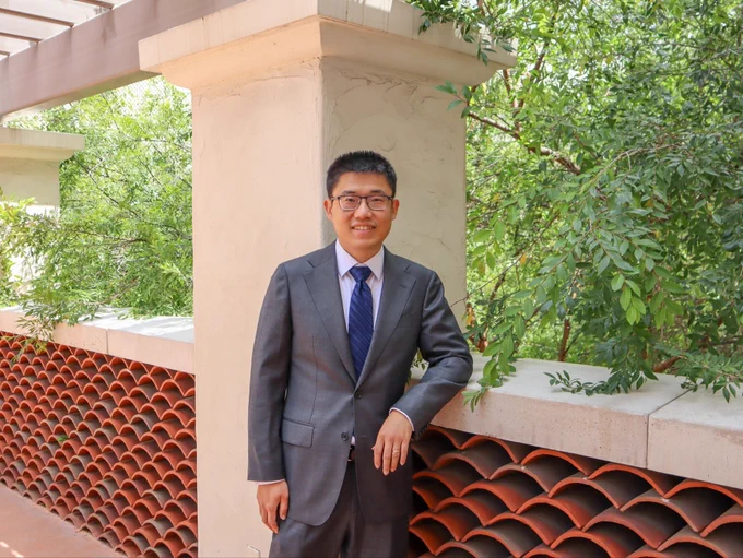 A person in a suit stands on a balcony with a red-tiled barrier, surrounded by lush green trees.
