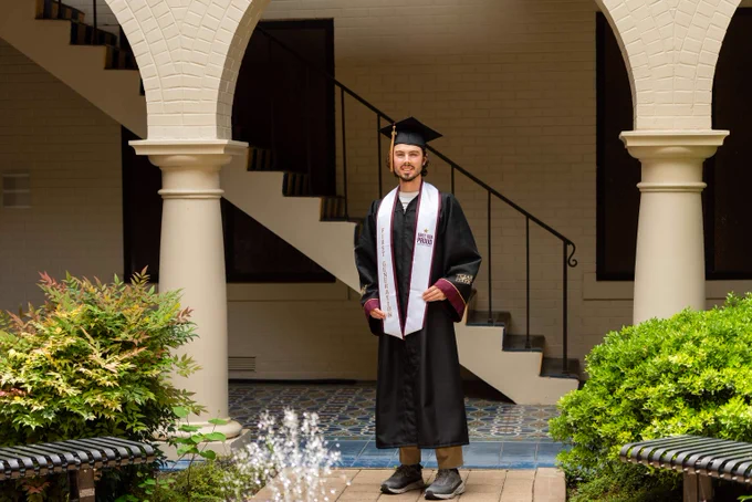 Graduate in black cap and gown standing under an archway with stairs in the background and greenery on both sides.
