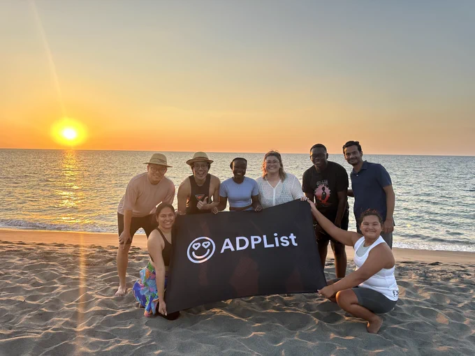 A group of eight people poses on the beach at sunset, holding an ADPList banner, celebrating a milestone together.