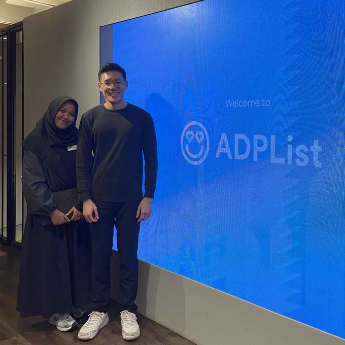 Two young adults stand together in front of a bright blue wall displaying “Welcome to ADPList.” The atmosphere feels welcoming and collaborative.