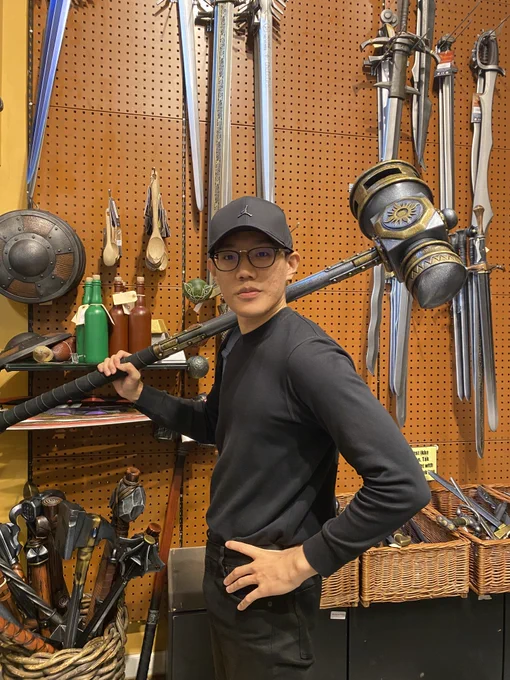 A person in casual attire poses confidently with a long staff, surrounded by various swords and weapons displayed on a wall.