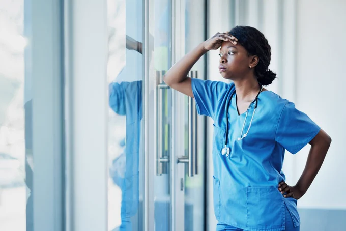 A healthcare worker stands and rests her elbow on a window. She places her hand on her forehead and looks out the window while exhaling. She appears to be stressed. 