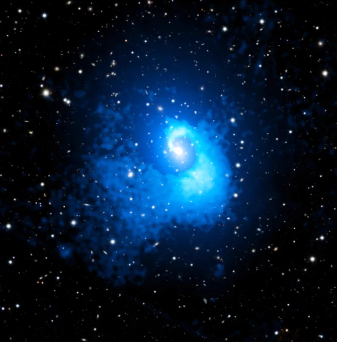 This is a composite image of galaxy cluster Abell 2029, a galaxy cluster with a unique spiral shape, giving it the appearance of a giant galactic seashell floating in the star-speckled blackness of space.

The surrounding stars and individual galaxies appear white, captured in optical light from Pan-STARRS, a telescope in Hawaii. But much of the spiraling cluster is rendered in neon blues, representing X-ray gas observed by Chandra. This super-heated gas fills the space between galaxies, giving the cluster its spiral shape when observed by scientists using an X-ray telescope.

Here, the blue spiral begins as a pale blue dot at the center of the cluster. The spiral stream of light and dark neon blue gas then widens as it moves away from the center of the cluster, gently corkscrewing one full rotation as it extends two-million lightyears into the distance.