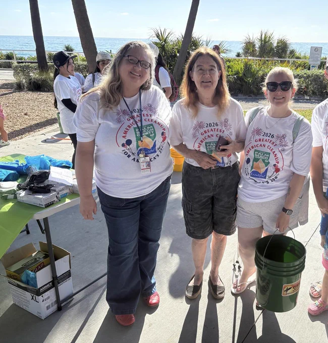 Three volunteers wearing matching 2026 American Cleanup event t-shirts stand outdoors near the beach with cleanup supplies nearby.