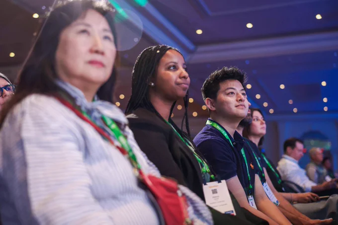 Audience members wearing conference badges sit attentively in a dimly lit event hall, facing a stage off-camera.