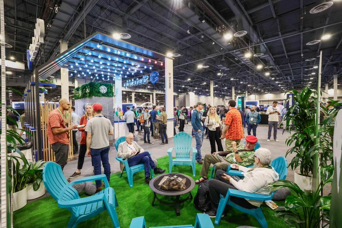 Busy trade show floor with a lounge area of blue chairs on green turf, surrounded by plants and people talking under bright lights.