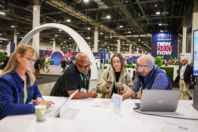Four attendees talk around a table with laptops and phones at a busy convention hall with a “new next now” banner.