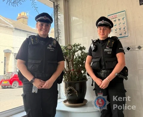 Two female police officers in uniform stand indoors, with a plant behind them.
