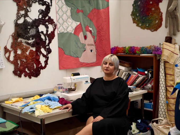A portrait of Ainaz Alipour seated in their textile studio beside a sewing machine and fabric materials. They are wearing a black dress and sitting in front of colorful textile artworks and sculptural pieces hanging on the walls, surrounded by books, tools, and works in progress.
