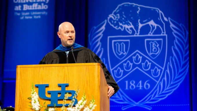 At a podium is, Joseph Stefko, a speaker wearing graduation attire. Behind them is the University at Buffalo logo with the text \"University at Buffalo The State University of New York\". The university logo has text reading \"1846\" at the bottom.