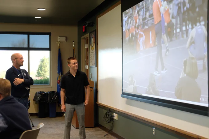 Two men watch a wrestling match projected on a screen in a classroom setting with chairs and a window in the background.