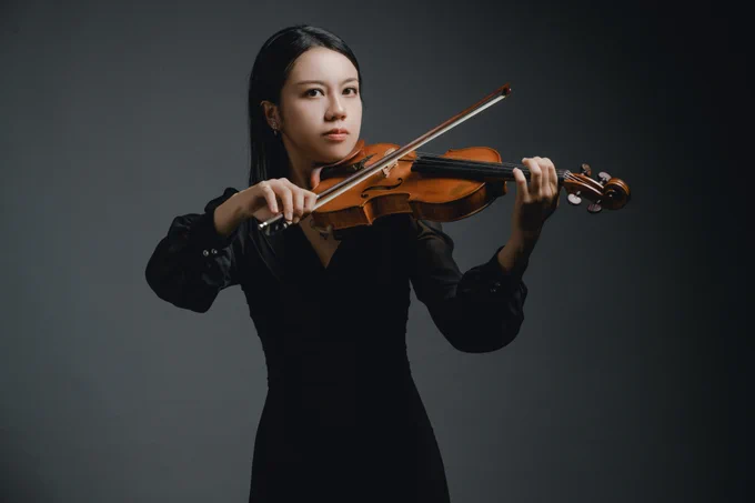 A young woman with long dark hair and a focused expression plays a wooden violin. She wears all-black performance attire and is photographed against a neutral gray background in a professional headshot-style composition.