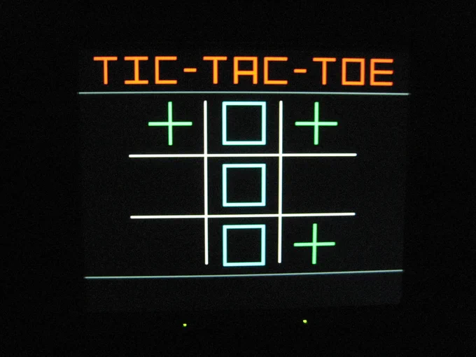 A digital display of a Tic-Tac-Toe game. The board shows two empty squares in the first column, three blue squares filled with blue Xs in the middle column, and two green plus signs in the third row. The words "TIC-TAC-TOE" are displayed at the top.