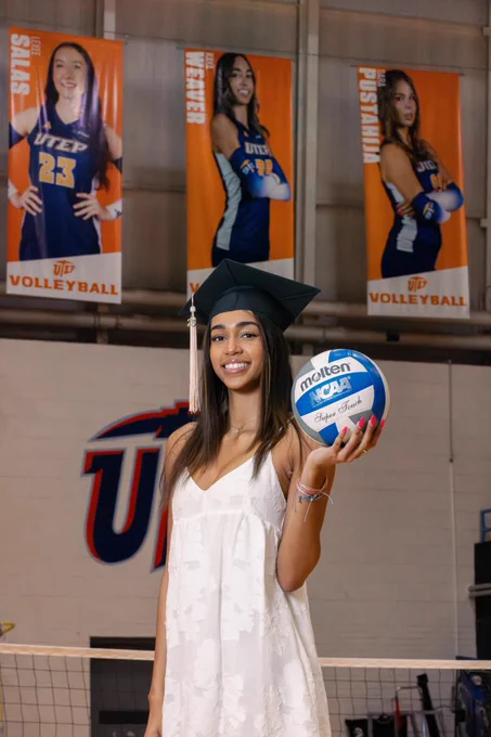Kaya Weaver in a white dress and graduation cap stands holding a volleyball. Banners featuring UTEP volleyball players hang in the background, as well as a volleyball net. 
