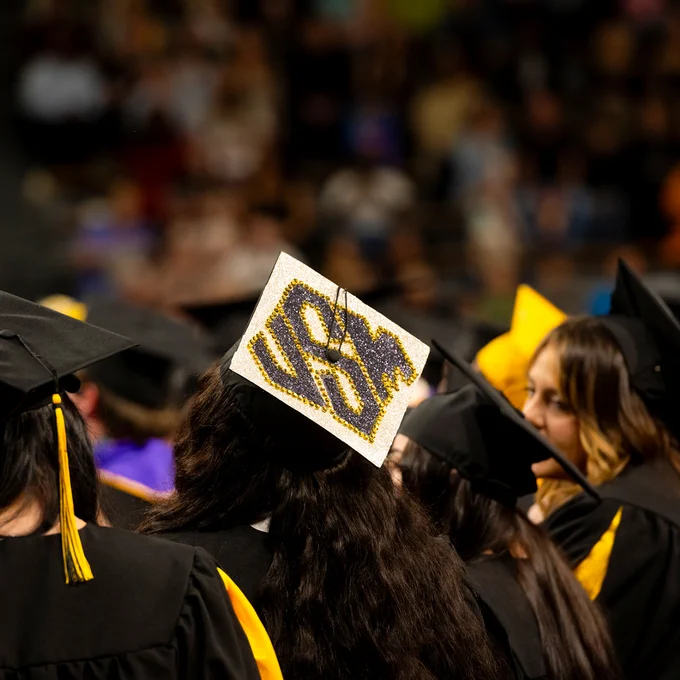 From behind, a graduation cap decorated in black and gold rhinestones reading "USM."