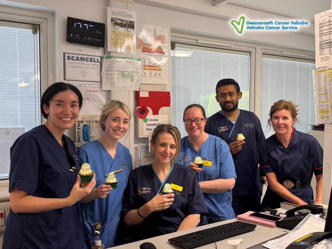 Five healthcare workers in navy and blue scrubs holding cupcakes inside a clinical office.
