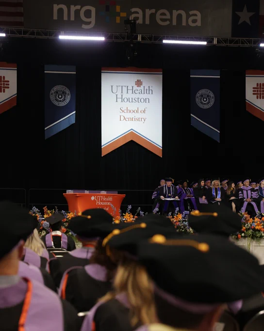 A graduation ceremony takes place on a large indoor stage where faculty in purple and black academic regalia are seated and standing. Banners for UTHealth Houston School of Dentistry hang behind them, and the stage is decorated with orange and purple flowers. Rows of graduates sit facing the stage.