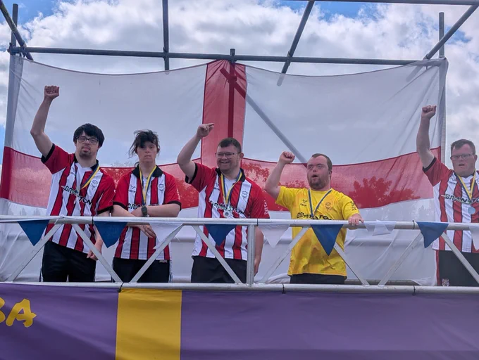 Group of young fans wearing red and white striped football jerseys raising fists in front of a large England flag outdoors.