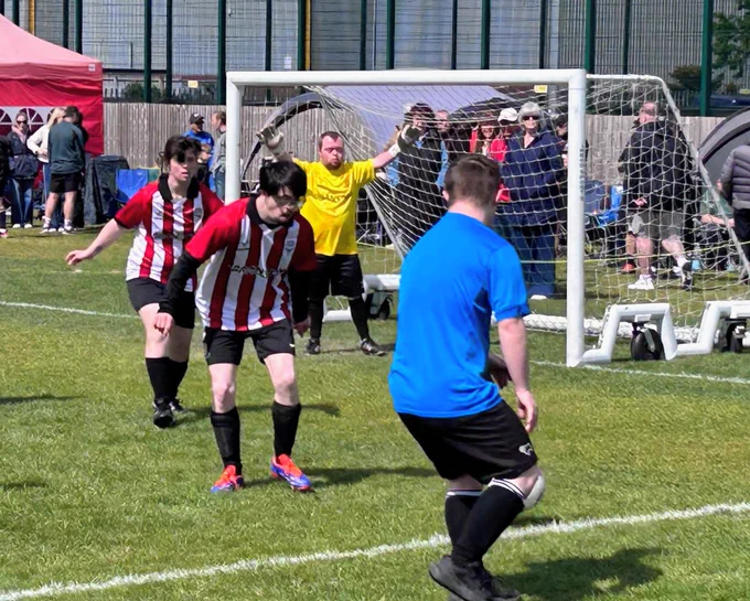 Players in red and white stripes defend against a player in blue near the goal. The goalkeeper, in yellow, watches intently. Spectators are in the background.