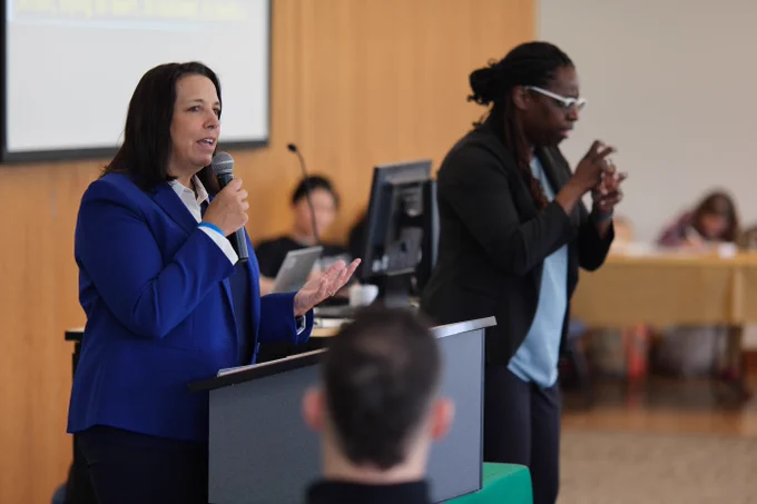 Lieutenant Governor Kim Driscoll delivers speech at Holyoke Community College beside interpreter for MCDHH 40th anniversary event. She is wearing a blue blazer and white collared shirt.