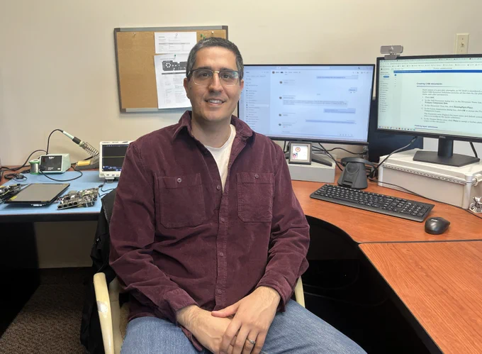 New Phason Controls employee Saleh Ebrahimi seated at a workstation with computer monitors and electronics equipment highlighting his role in hardware and embedded systems development.
