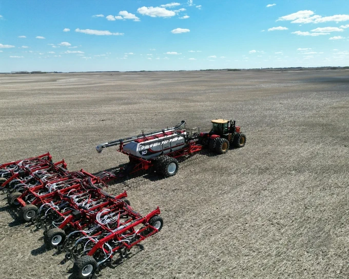 A large Morris 10 series air cart is being towed across a vast, empty field, followed by a Quantum seeder. The sky is clear with a few scattered clouds on a bright sunny day.