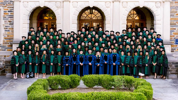 A large group of graduates (MD and MD/PhD) wearing green academic gowns and caps stand in several rows in front of a stone building with arched windows. Four individuals in blue robes are seated in the front row. The scene takes place outdoors, with a small hedge in the foreground.