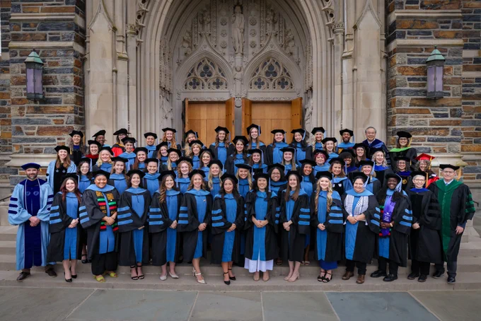 A group of graduates (Occupational Therapy Doctorate graduates) in robes and academic regalia pose in front of a stone building with ornate wooden doors. Some wear blue attire, and a few are in green or have colorful stoles. The setting has a formal, celebratory atmosphere.
