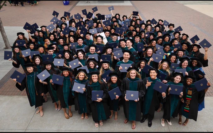 A large group of graduates  (Doctor of Physical Therapy graduates) in green robes proudly hold their diplomas, standing outdoors on a paved area. They are smiling and looking up towards the camera.