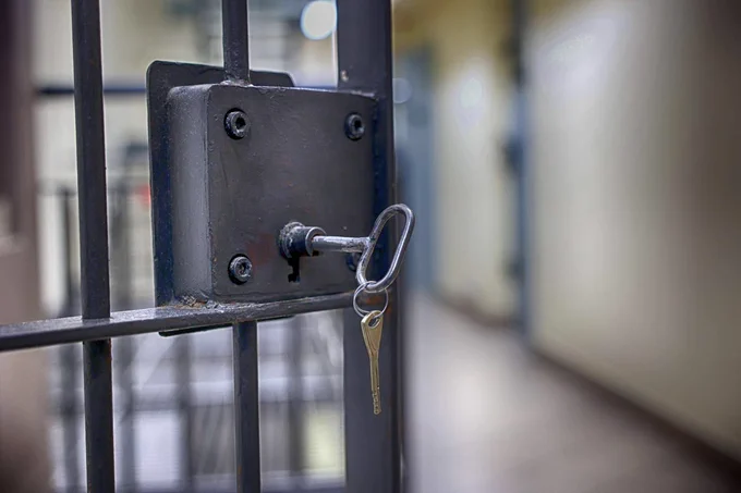 Close-up of a metal lock on a prison cell door with a key inserted, showing the bars and blurred corridor background.