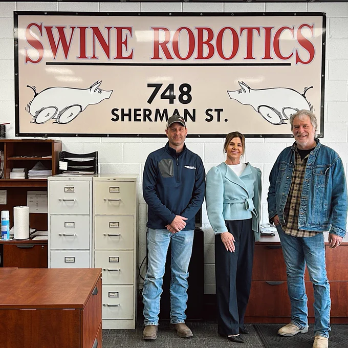 Three individuals stand in an office at Swine Robotics, located at 748 Sherman St. Behind them, a sign with pigs in motion is displayed. They are positioned in front of filing cabinets and desks.