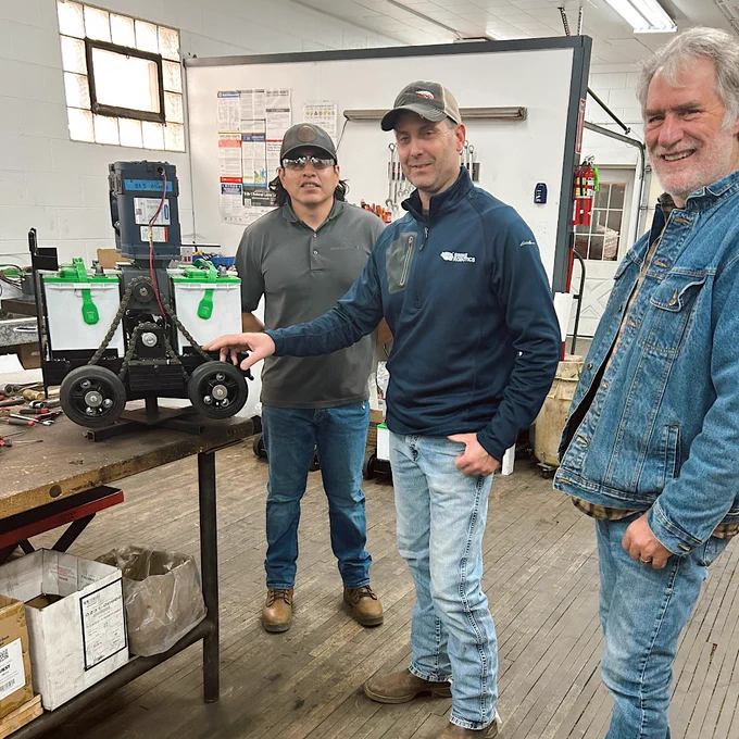 A group of three people stand in a workshop. They are next to a wheeled device resembling a small vehicle. The room contains tools, shelves, and informational posters on the walls.